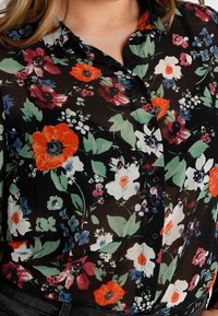 Chemise noire à boutons ornée d'un motif floral vibrant avec des fleurs rouges, blanches, bleues et roses, ainsi que des feuilles vertes, portée par une personne.