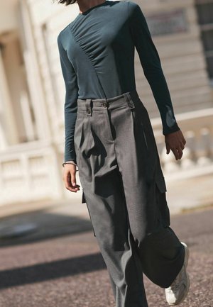 Maglia a maniche lunghe di color teal con dettaglio arricciato sul davanti, abbinata a pantaloni cargo grigi a vita alta con doppio bottone e tasche laterali.
