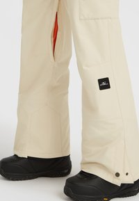 Beige waterdichte broek met ritsen aan de zijkanten en een zwart O'Neill-logo, gecombineerd met zwarte, gestructureerde sneeuwlaarzen met een robuuste zool.
