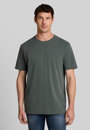Man met kort, krullend bruin haar, die een effen donkergroen T-shirt met korte mouwen en een blauwe spijkerbroek draagt tegen een lichtgrijze achtergrond.