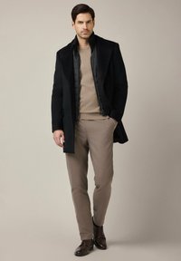 Czarny długi płaszcz na pikowanym kamizelce, beżowy sweter i jasnobrązowe spodnie. Brązowe skórzane buty. Prosty, nowoczesny design o dopasowanym kroju.