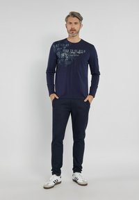 Granatowa koszula z długim rękawem z nadrukiem graficznym, w połączeniu z ciemnymi spodniami w paski i białymi trampkami. Luźny krój, gładki materiał, ribbowane mankiety.