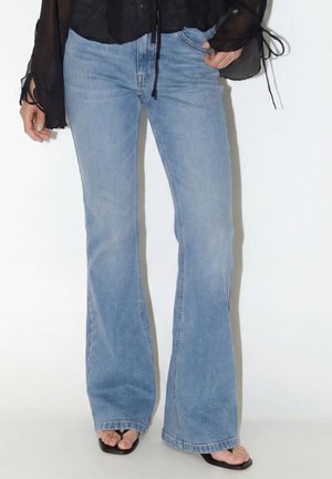 Vrouw draagt lichtblauwe wijde jeans met zwarte sandalen met hak en een semi-transparante zwarte blouse met gestrikte manchetten.