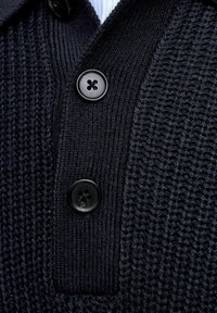 Nahaufnahme eines dunkelblauen Strickpullis mit zwei großen schwarzen Knöpfen, die an der gerippten Leiste und dem strukturierten Strickstoff befestigt sind.