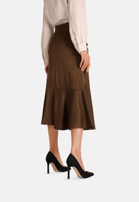 Gonna in satin marrone con orlo svasato, abbinata a scarpe con tacco alto nere. La gonna presenta una vita aderente e una texture liscia.