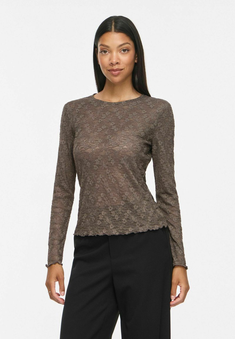 Femme aux longs cheveux noirs portant un haut en dentelle marron ajusté à manches longues et un pantalon noir, debout devant un fond blanc uni.