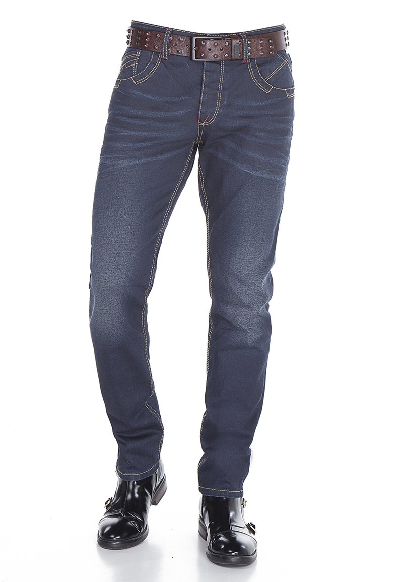 Donkerblauwe denim jeans met contrasterende gele stiksels, vijfzakken ontwerp en een bruin gesp-studded riem in de taille. Zwarte enkelboots eronder.