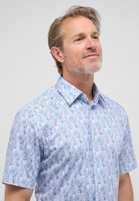 Korte mouw shirt van lichtblauwe stof met een bloemenpatroon met blauwe en groene bladeren. Klassieke kraag en knopen voor sluiting.
