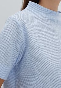 Top azzurro chiaro caratterizzato da un tessuto testurizzato e rigato, con colletto alto e maniche corte. Il design enfatizza comfort e semplicità.