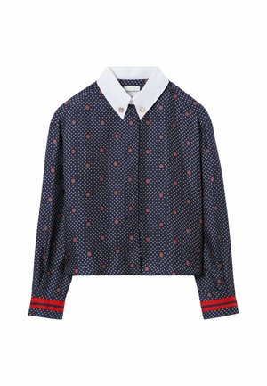 Personne debout portant une chemise bleu marine à motifs avec col blanc, pantalon beige à jambes larges, pull jaune noué à la taille et chaussures marron.