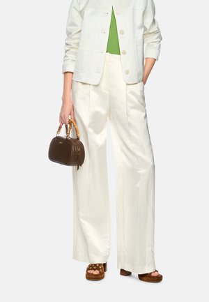 Woman wearing white wide-leg pants, white jacket, green top, brown heeled sandals, and holding a small brown handbag with bamboo handles.