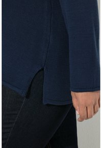 Maglione a maglia blu navy con texture a costine e spacchi laterali, abbinato a jeans scuri. Il tessuto appare morbido e flessibile.