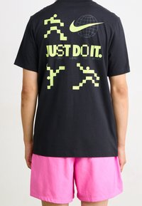 Musta t-paita, jossa on keltaisia pikselöityjä juoksuhahmoja, Nike-logon päällä maapallo ja teksti "JUST DO IT." yhdistettynä vaaleanpunaisiin shortseihin.