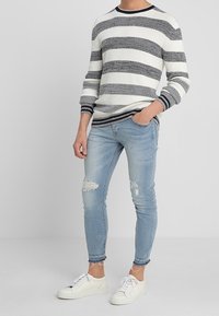 Raidallinen neulepusero valkoisena ja laivastonsinisenä, yhdistettynä vaaleansinisiin skinny-farkkuihin, joissa on repeytyneet yksityiskohdat ja purjehdushelmat, viimeistelty valkoisilla lenkkareilla.