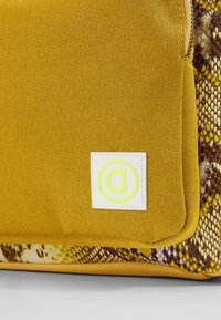 Bolso texturizado amarillo con un acento de patrón de serpiente. Presenta una etiqueta blanca con un logo verde. Cierre con cremallera y bordes reforzados.