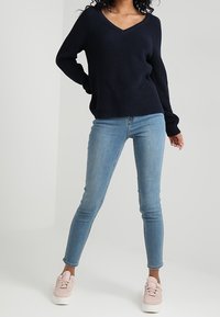 Pull en tricot bleu marine à col en V, associé à un jean skinny bleu clair et des baskets rose pâle, présentant une tenue décontractée.