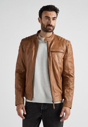 Veste en cuir brun avec fermeture éclair à l'avant, détails côtelés aux épaules et poches latérales. Portée sur un pull gris clair et un jean sombre.