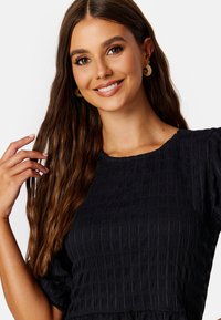 Zwarte top met textuur, voorzien van een smockontwerp, korte pofmouwen en een ronde halslijn. Het model heeft lang, golvend haar en draagt oorbellen.