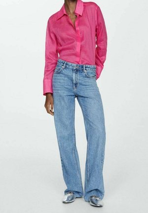 Person iført en knaldpink langærmet bluse stukket ned i løstsiddende lyseblå jeans med sølvfarvede spidse sko mod en ensfarvet baggrund.