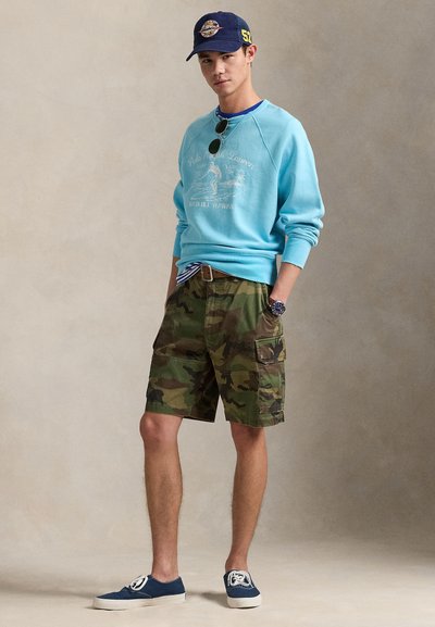 Ανοιχτό μπλε φούτερ με γραφικό τύπωμα, σορτς παραλλαγής, μπλε αθλητικά παπούτσια και μπλε σκούφος. Αξεσουάρ περιλαμβάνουν γυαλιά ηλίου και ζώνη με ρίγες.