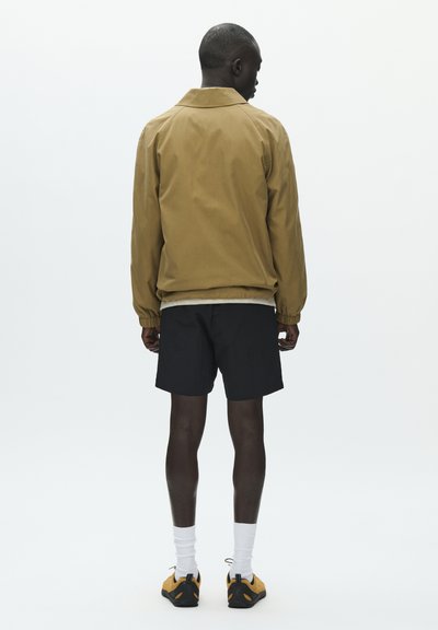 Homme vu de dos portant une veste beige, un short noir, des chaussettes blanches et des baskets jaunes sur un fond blanc uni.