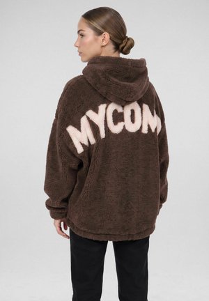 Vrouw met haar in een knot, die een bruine, zachte jas met capuchon draagt met grote witte letters die "MYCOM" op de achterkant spellen, zijwaarts staand.