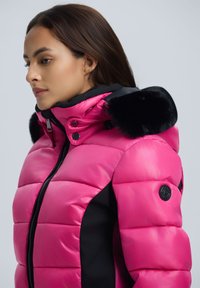 Chaqueta puffer rosa con un acabado brillante, paneles laterales negros, capucha con borde de pelo, cierres de cremallera y botón a presión, y un logo redondo en la manga.