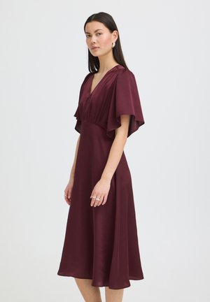 Kobieta o prostych, ciemnych włosach nosi sukienkę midi z długim rękawem w głębokim bordowym kolorze z dekoltem w serek na tle jednolitego, szarego tła.