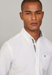 Camisa blanca de botones con cuello que presenta rayas azules y amarillas. Incluye un pequeño logo bordado en el bolsillo del pecho. Textura suave.