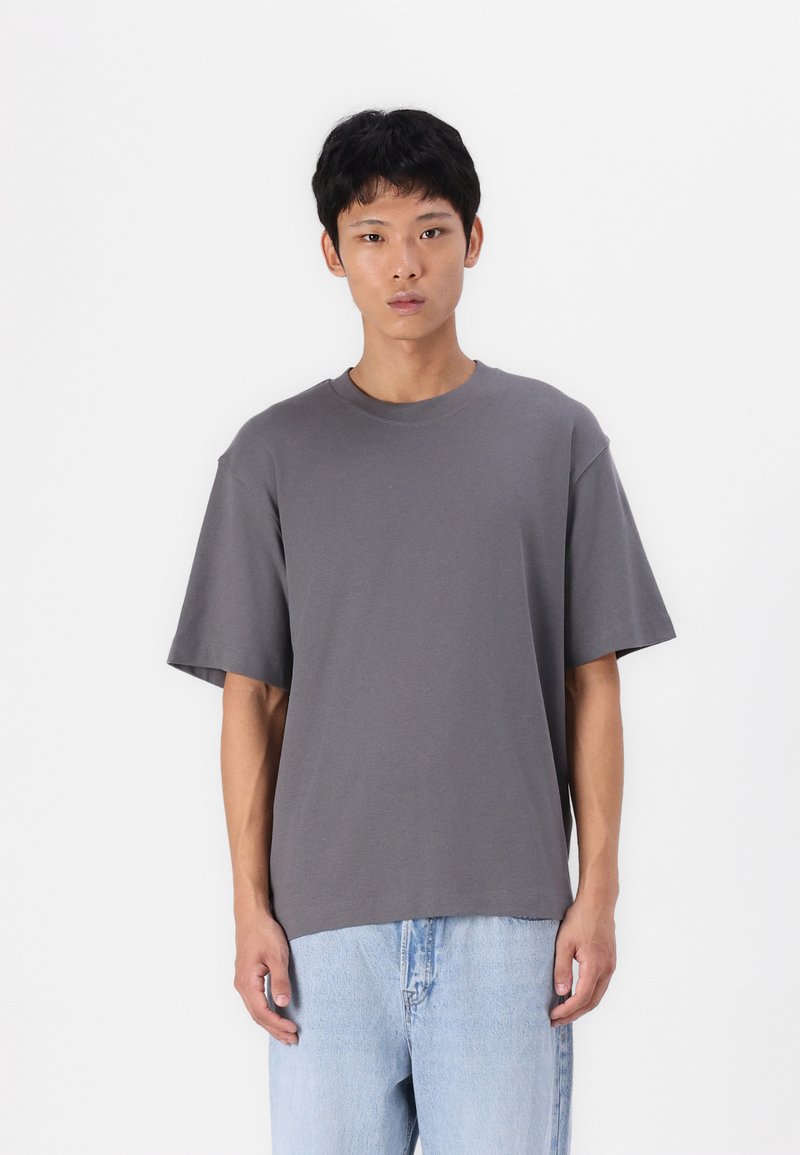 Tricou gri cu guler rotund, mâneci scurte, croială lejeră, fabricat dintr-un amestec de bumbac, asortat cu blugi din denim de culoare albastră deschisă.