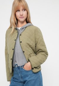 Pikowana oliwkowo-zielona kurtka z czarnymi zatrzaskami, okrągłym kołnierzem i bocznymi kieszeniami, założona na szary hoodie, w zestawieniu z jasnoniebieskimi dżinsami.