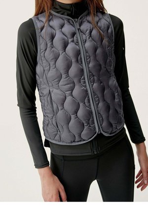 Kobieta nosząca czarną dopasowaną bluzkę z długim rękawem i czarną pikowaną kamizelkę bez rękawów z zamkiem z przodu.