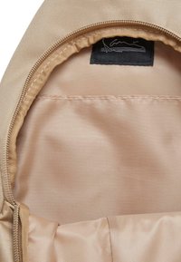 Beżowe wnętrze plecaka z gładką podszewką, z czarną metką logo i zamkiem błyskawicznym wzdłuż górnej krawędzi.