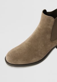 Brown suede ankle boot with a smooth texture, elastic sides, and a rounded toe. Features a low black sole with a subtle stitching detail.