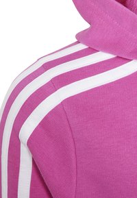 Sudadera rosa con tejido texturizado, presenta acentos de tres rayas blancas a lo largo de las mangas y una capucha con cuerda.