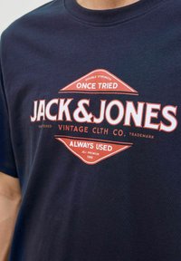 Tamsiai mėlynas medvilninis marškinėliai su raudonu ir baltu grafiniu spaudiniu, ant kurio užrašyta "JACK & JONES" bei papildomas tekstas "ONCE TRIED" ir "ALWAYS USED."