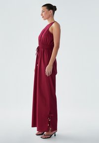 Bordo maxi obleka s poudarjenim pasom, brez rokavov, stranskimi razporki z zavezovanji in zlatimi poudarki. Gladek material z tekočo silhueto.