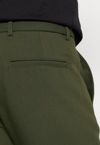 Pantaloni verde oliva con una texture liscia, dotati di una tasca posteriore con un pattina rettangolare e dettagli di cucitura sottili.