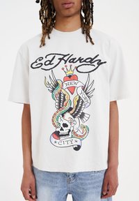 Camiseta blanca de algodón con un colorido estampado gráfico de un cráneo, un águila, corazones y serpientes, junto con el texto "Ed Hardy," "NUEVA YORK" y "CIUDAD."