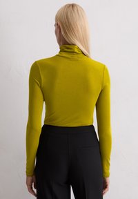 Langärmliger Rollkragenpullover aus leuchtend grünen Stoff, enganliegendes Design, kombiniert mit hochtaillierten schwarzen Hosen mit einer Rückentasche.