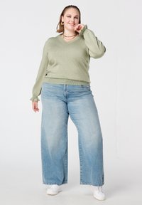 Lichtgroene V-hals trui met omgeslagen mouwen gecombineerd met lichtblauwe jeans met wijde pijpen. Model draagt witte sneakers en gelaagde metalen kettingen.