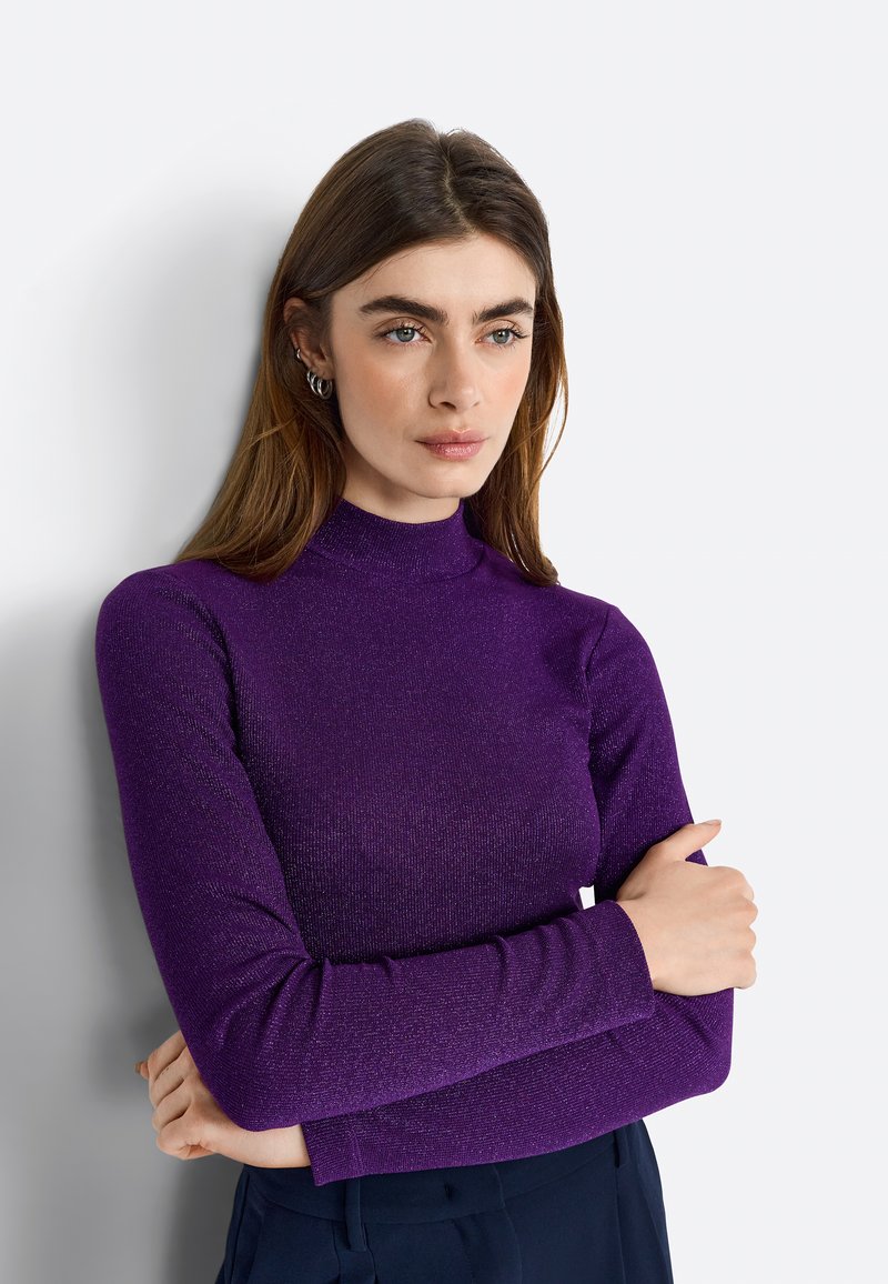 Top lung cu mâneci lungi de culoare purpurie, cu un gât înalt, fabricat dintr-un material texturat și strălucitor. Modelul își încrucișează brațele, purtând accesorii elegante, pe un fundal neutru.