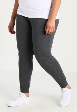 Person iført mørkegrå tætsiddende joggingbukser og hvide sneakers, der står på en lys baggrund.