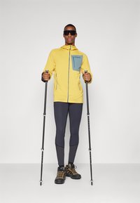 Yellow hooded jacket with a teal chest pocket, paired with navy leggings and black shoes, holding trekking poles, standing on a neutral background.