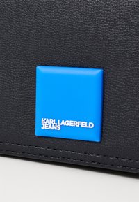 Material texturat negru cu un logo pătrat albastru, având inscripția "KARL LAGERFELD JEANS" în litere albe. Accente cu finisaj neted.