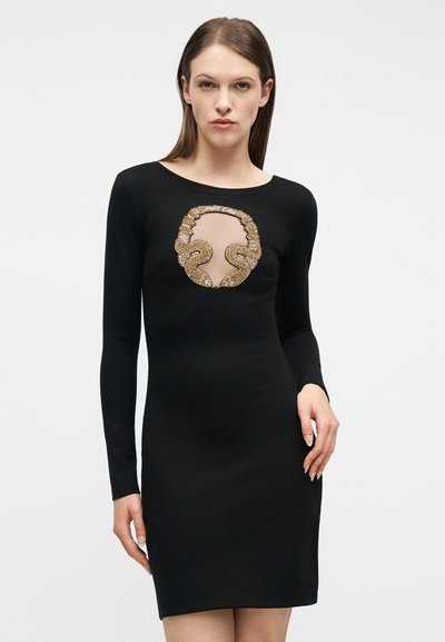 Γυναίκα με μαύρο φόρεμα μακρυμάνικο με διαφανές τμήμα στο στήθος και χρυσή παγιετέ φιδίσια σχεδίαση, που στέκεται μπροστά σε απλό λευκό φόντο.