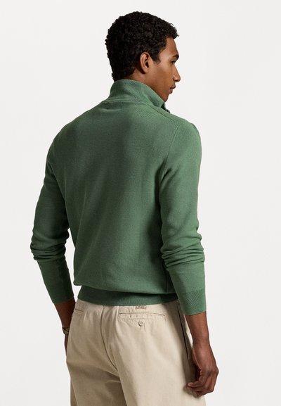 Green knit pullover sweater with a high collar, textured pattern, and fitted cuffs, paired with light beige pants.