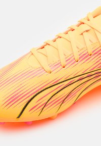 Botín de fútbol amarillo brillante con rayas diagonales rosas, acentos negros y superficie texturizada; cuenta con cordones estándar y clavos redondeados.