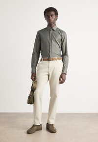 Olivgrünes Hemd mit blauen Knöpfen, cremefarbene Hose, tanfarbener Gürtel und braune Sneaker, hält eine grüne Tasche mit verstellbarem Tragegurt.
