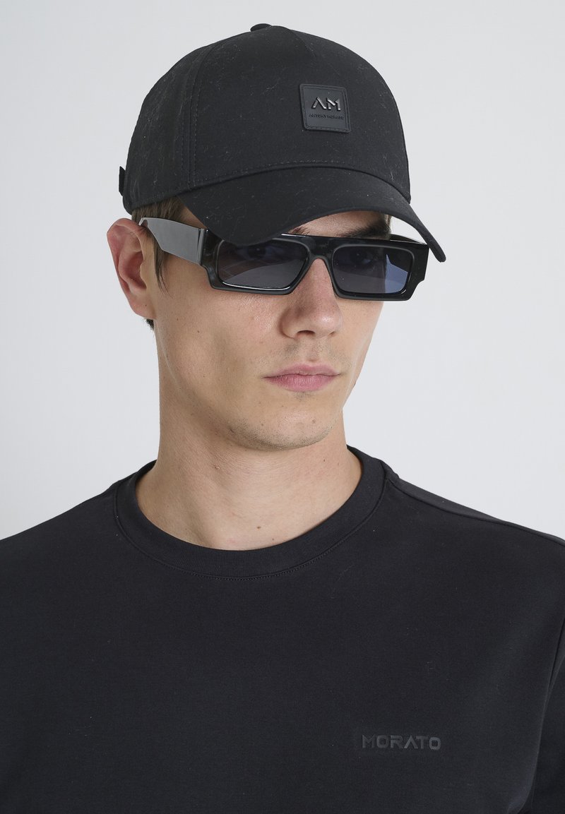 Gorra negra con un logotipo, gafas de sol rectangulares negras y una camiseta negra con un sutil detalle de marca en el pecho.